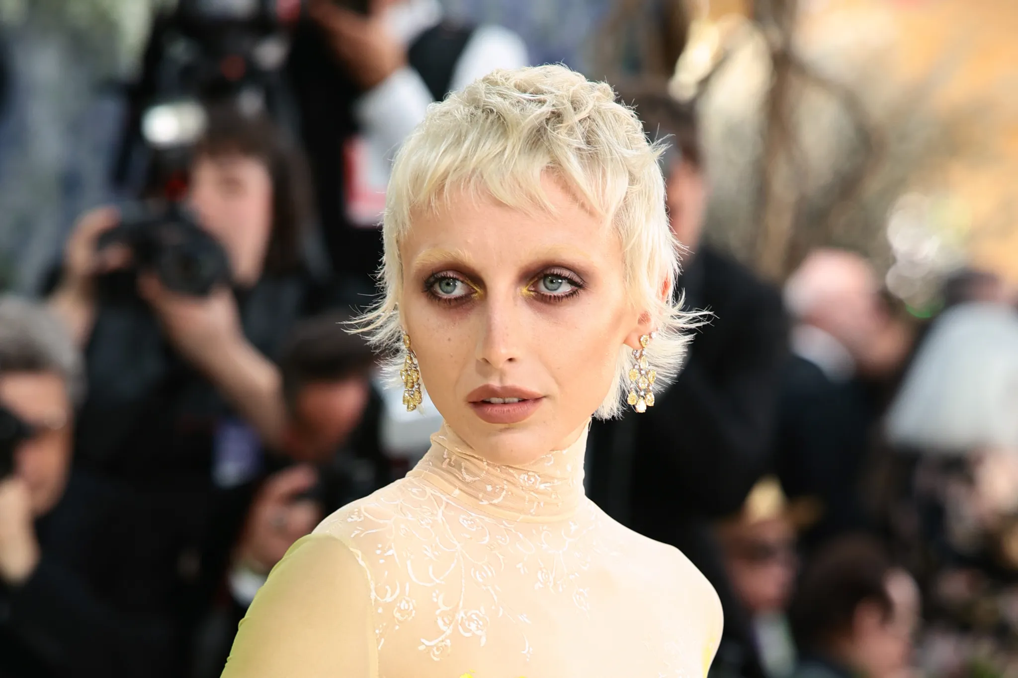 Met Gala 2026: Emma Chamberlain, redaktorka časopisu Vogue, na červeném koberci zvolila výrazný žlutý oční stín ve vnitřním koutku oka, aby ladil s její ručně malovanou zakázkovou róbou od Muglera.